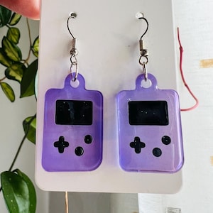 May include: A pair of drop earrings featuring purple, translucent, rectangular pendants shaped like handheld video game consoles. Each pendant has a black screen area and black circular and cross-shaped buttons. The earrings hang from silver-toned metal hooks.