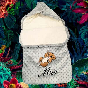 Peut inclure: Une gigoteuse bébé grise avec une doublure blanche et un petit lion brodé sur le devant. La gigoteuse est à pois gris et le nom "Mio" est brodé sous le lion.