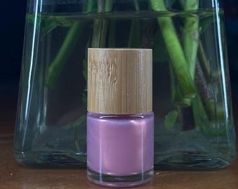 Può includere: Un piccolo flacone di smalto per unghie rosa con un tappo in legno. Il flacone è appoggiato su una superficie di legno.
