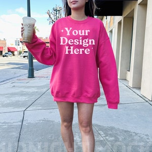 Puede incluir: Una sudadera rosa brillante con el texto "Your Design Here" en blanco. La sudadera de cuello redondo se usa con zapatillas blancas. La imagen est&aacute; tomada al aire libre en una acera.