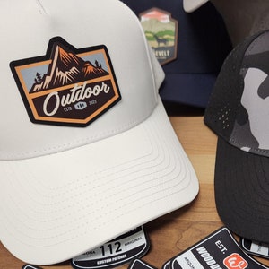 May include: Two baseball caps with embroidered patches. The white cap has a brown, orange, and blue patch with the text "Outdoor" and "Est. 2021". The black cap has a camouflage pattern and a brown, orange, and blue patch with the text "The Mountain Exploration Est. 2023".