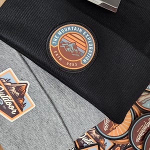 May include: Close-up of two beanies, one black and one gray, with mountain-themed patches. The black beanie has a patch that reads "The Mountain Exploration Est. 2023." The gray beanie has an "Outdoor" patch. Additional mountain-themed stickers are visible.