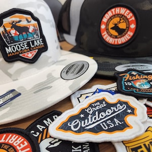May include: A collection of trucker hats and patches with outdoor themes. The hats feature patches with designs like a moose in a landscape, and a deer with the text "Northwest Territories". Patches include "The Great Outdoors U.S.A."
