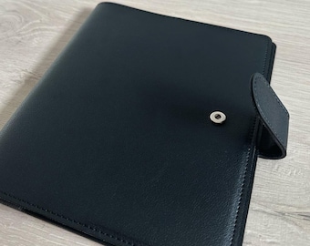 May include: A black leather portfolio with a snap closure. The portfolio is rectangular and has a smooth texture. The closure is a small, silver-colored snap. The portfolio is closed and appears to be empty.