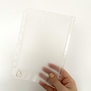 Könnte beinhalten: Transparente Plastik-Schutzfolie mit Reißverschluss und Löchern für einen Ordner. Die Schutzfolie hat ein kleines Wassertropfen-Logo in der linken unteren Ecke.