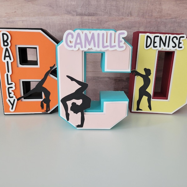 May include: Three-dimensional decorative letters spelling out names, with a gymnastics theme. The letters are orange, pink, and yellow, each with a black gymnast silhouette. The names Bailey, Camille, and Denise are displayed.