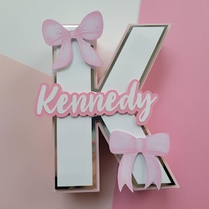 May include: A white letter K with a silver border, decorated with pink bows and the name "Kennedy" in pink letters.