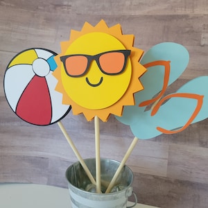 May include: A summer-themed centerpiece with a yellow sun wearing sunglasses, a red and white beach ball, and two blue flip-flops.