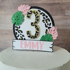 May include: A cake topper featuring a leopard print circle with a gold glitter number 3, surrounded by green and pink leaves. The topper also includes a white banner with the name "EMMY" in pink letters.