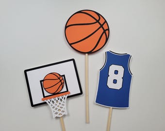 Puede incluir: Tres adornos para pasteles con temática de baloncesto. Un adorno presenta una pelota de baloncesto naranja con líneas negras. Otro adorno presenta una camiseta de baloncesto azul con el número 8 en la parte delantera. El tercer adorno presenta un aro de baloncesto con una red y una pelota de baloncesto naranja en su interior.