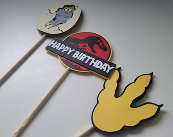 May include: Three dinosaur-themed cake toppers on wooden sticks. One shows a baby dinosaur hatching from an egg, another has a red and black logo with "HAPPY BIRTHDAY", and the last is a yellow dinosaur footprint.