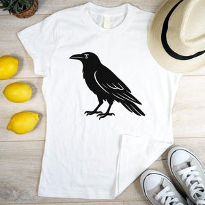 May include: White t-shirt featuring a black raven graphic. The raven is depicted in a simple, stylized design. The shirt is on a wooden surface with lemons, a hat, and sneakers.