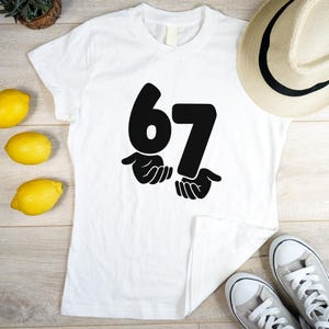 Puede incluir: Camiseta blanca con un gráfico negro del número 67, sostenido por manos. La camiseta está sobre una superficie de madera, con limones, un sombrero y zapatillas en el fondo.