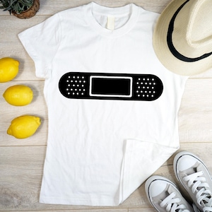 Pode incluir: Camiseta branca com um desenho de penso preto. O penso tem um centro retangular e bordas pontilhadas. A camiseta está em uma superfície de madeira com limões, um chapéu e tênis.