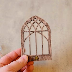 May include: A wooden night light shaped like a cathedral window. The light has a detailed arched frame with a white interior and a small brass-colored button at the bottom. The night light is held in a person's hand.