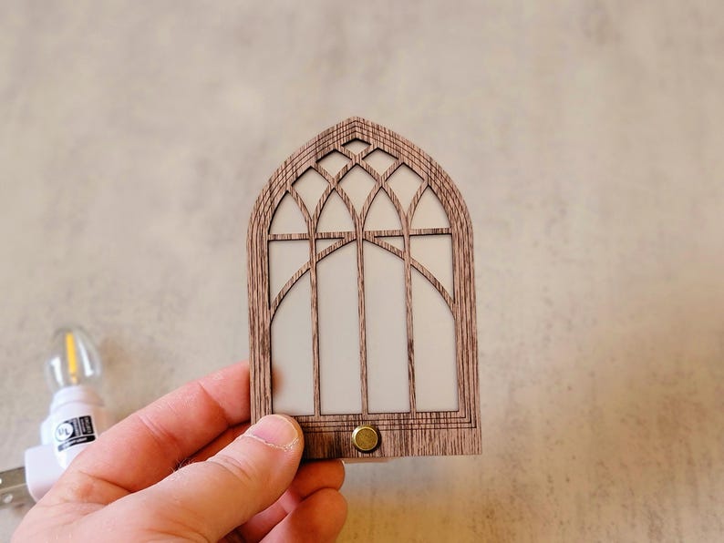 May include: A wooden night light shaped like a cathedral window. The light has a detailed arched frame with a white interior and a small brass-colored button at the bottom. The night light is held in a person's hand.