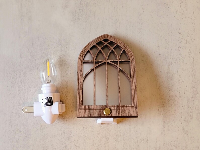 May include: A decorative night light featuring a wooden arched window design with a warm, inviting glow. The night light has a white base and a small, energy-efficient bulb. The design is reminiscent of a church window.