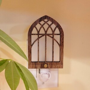 May include: Wooden night light shaped like a cathedral window. The frame is a dark brown color, with a white interior. The night light is plugged into a white outlet. The design is reminiscent of a church window.