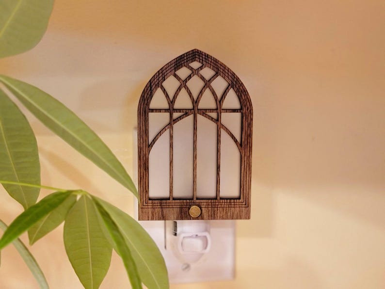 May include: Wooden night light shaped like a cathedral window. The frame is a dark brown color, with a white interior. The night light is plugged into a white outlet. The design is reminiscent of a church window.