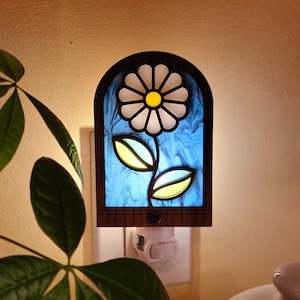 Puede incluir: Una luz nocturna con forma de vitral con fondo azul y una margarita blanca con centro amarillo. La luz nocturna está enchufada a una toma de corriente.