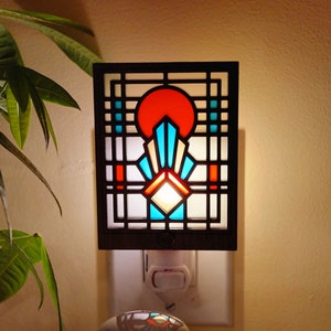 May include: A stained glass night light with a geometric design in shades of red, blue, and white. The light is plugged into an outlet and is illuminated.