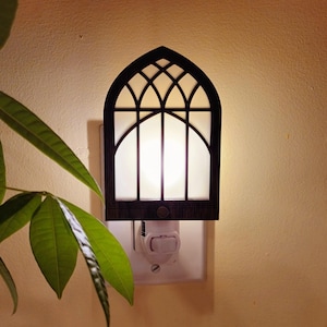 May include: A decorative night light with a dark brown, arched frame resembling a church window. The light has a white, glowing center and is plugged into a wall outlet. Green leaves from a nearby plant are visible in the foreground.