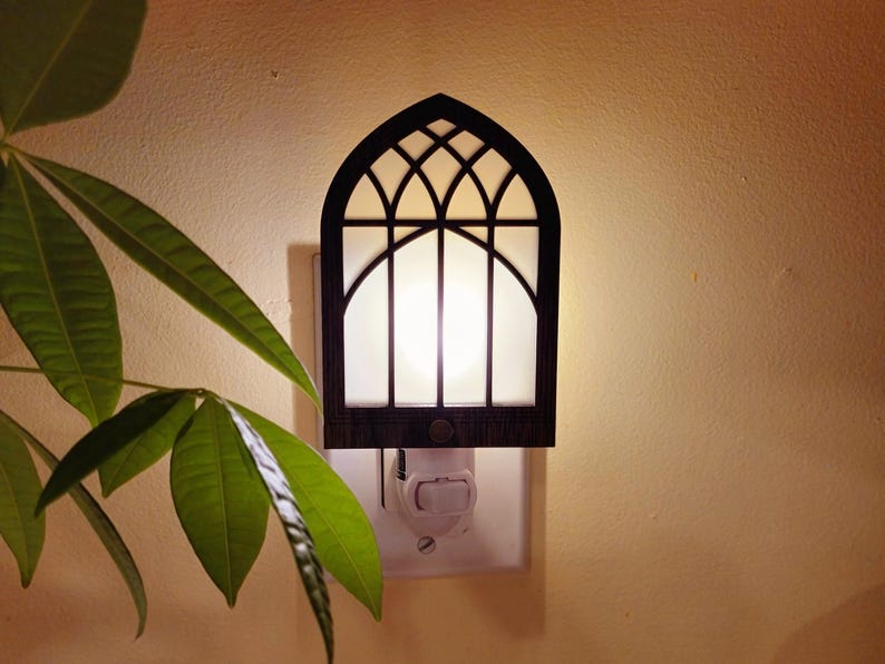 May include: A decorative night light with a dark brown, arched frame resembling a church window. The light has a white, glowing center and is plugged into a wall outlet. Green leaves from a nearby plant are visible in the foreground.