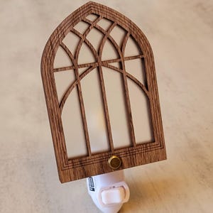 May include: Wooden night light with a cathedral window design. The light has a brown wooden frame with a white interior and a brass-colored button. The night light plugs into a standard wall outlet.