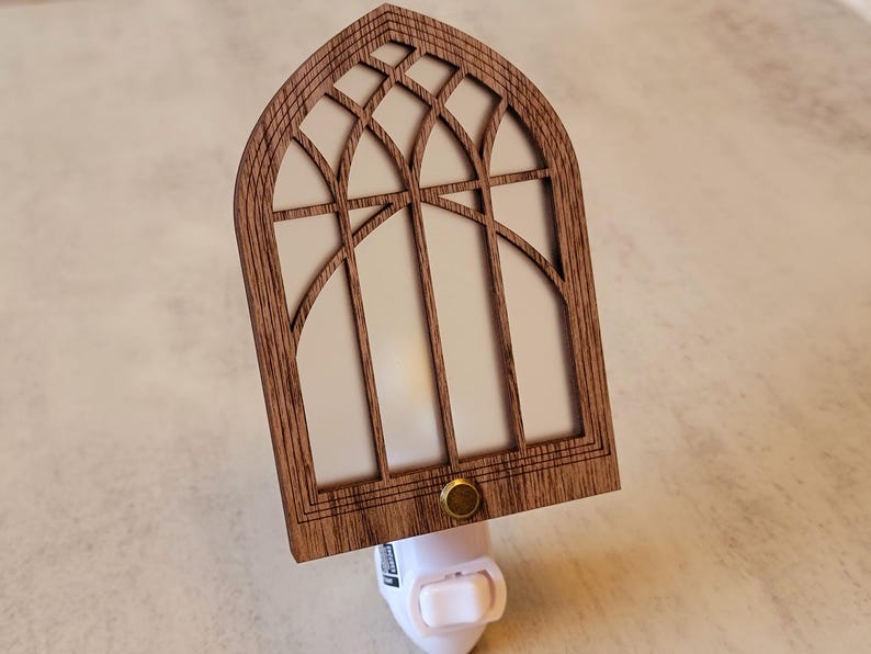 May include: Wooden night light with a cathedral window design. The light has a brown wooden frame with a white interior and a brass-colored button. The night light plugs into a standard wall outlet.