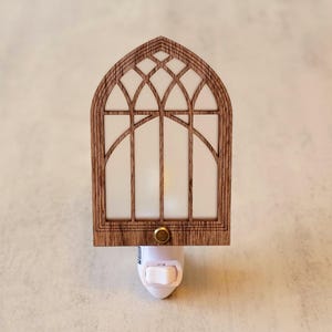May include: A wooden night light shaped like a cathedral window. The frame is a warm brown, with a white, translucent panel. The night light has a small, gold-colored button and a white plug.
