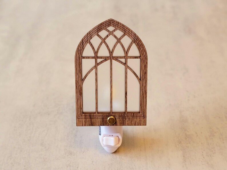May include: A wooden night light shaped like a cathedral window. The frame is a warm brown, with a white, translucent panel. The night light has a small, gold-colored button and a white plug.