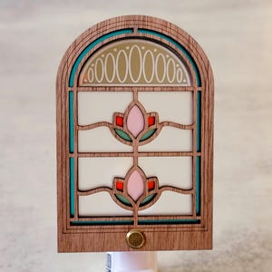 May include: A wooden nightlight shaped like a window with a teal frame and two pink flowers. The window has a white background and a decorative arch with gold ovals.