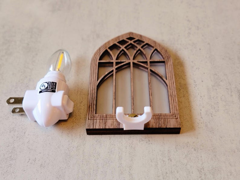 May include: A white night light with a UL label next to a wooden decorative window frame with a white holder. The window frame has a gothic arch design.