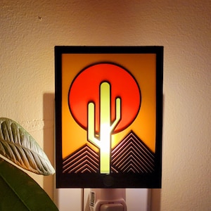 May include: A decorative night light featuring a desert landscape design. The image shows a black-framed square with a red sun, a green cactus, and geometric mountain shapes. The light is plugged into a wall outlet.