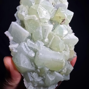 May include: A cluster of pale green, translucent crystals with a geometric, angular structure. The crystals are tightly packed together, creating a textured surface. The overall shape is irregular, with varying sizes and shapes of crystals. The background is black.