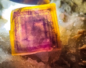 May include: A close-up of a purple and yellow fluorite crystal with a square shape. The crystal is sitting on a white, sparkly surface.