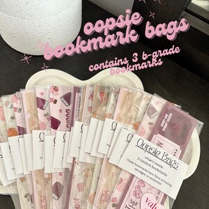 Puede incluir: Un conjunto de 16 bolsas de marcadores de libros rosas y blancas, cada una con tres marcadores de libros. Las bolsas están etiquetadas con "oopsie bookmark bags" y "what's inside: 3 random bookmarks". Las bolsas están dispuestas en círculo sobre una superficie blanca.