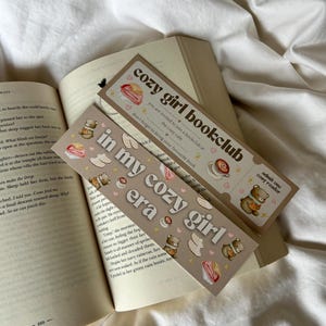 Peut inclure: Deux marque-pages marrons avec des illustrations d'ours, de pâtisseries et de cœurs. Le texte sur les marque-pages est "Cozy Girl Bookclub" et "In My Cozy Girl Era".