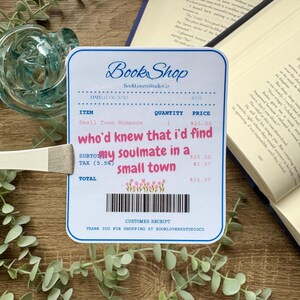 Op de afbeelding: Een bon van een boekwinkel met de tekst "Book Shop" en details van een aankoop van "Small Town Romance". De bon bevat de roze tekst "who'd knew that I'd find my soulmate in a small town", een streepjescode en een bloemenontwerp.