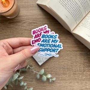 May include: A sticker with the text "BOOKS ARE MY EMO ARE MY EMOTIONAL SUPPORT" in pink and blue, with a small blue bow graphic. The sticker is held in a hand, with a lit candle and an open book in the background.