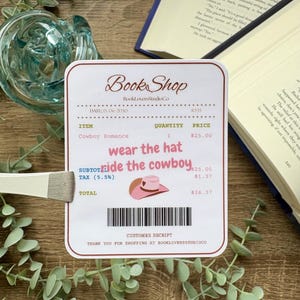 Op de afbeelding: Een sticker ontworpen als een kassabon van Book Shop. De bon bevat de tekst "Cowboy Romance", "wear the hat, ride the cowboy" en een afbeelding van een roze cowboyhoed. Het totaalbedrag is $26,37.