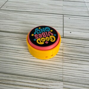 May include: A round, yellow plastic button with a pink middle layer and a black top surface. The black surface features the text "GOOD VIBES ONLY" in yellow, stylized lettering, accented by small blue and pink star-shaped icons.