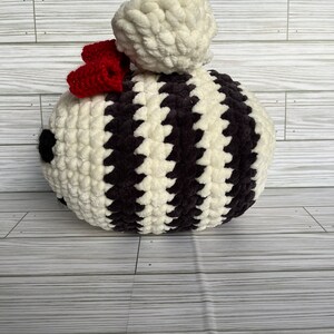 May include: A handmade, round, plush decorative item with alternating cream and dark brown stripes. A small red bow is attached to the side, and a large cream-colored pom-pom sits on top. The item is likely a decorative pillow or stuffed toy.