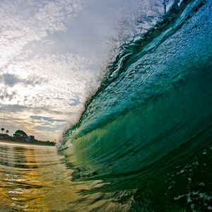 May include: A wave breaking in the ocean, with a view of the sky and shoreline in the background. The wave is a vibrant green color, and the water is a golden color.