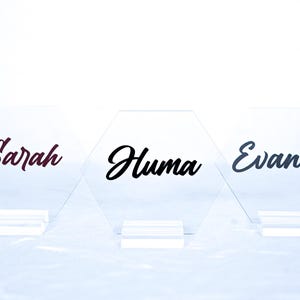 May include: Three clear acrylic hexagonal table cards with black and burgundy lettering. The names "Sarah", "Huma", and "Evandro" are written on the cards.