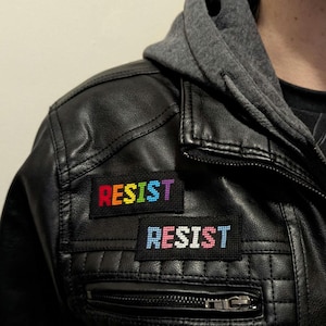 May include: A black leather jacket with two rainbow embroidered patches that say "RESIST" on them.