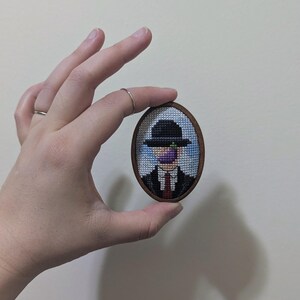 May include: A small, framed cross-stitch artwork depicting a man wearing a black hat and a purple fruit over his mouth. The artwork is framed in a brown oval frame.