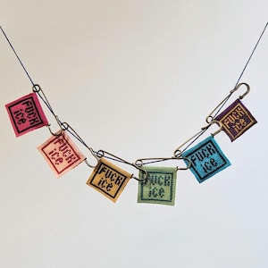 Peut inclure: Un collier fait à la main composé de six petits carrés brodés de différentes couleurs, chacun portant les mots "FUCK ICE" en noir. Les carrés sont reliés par des épingles de sûreté et suspendus à un fil bleu.
