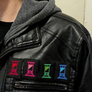 May include: Four embroidered patches sewn onto a black leather jacket. Each patch features a stylized hourglass design with a different color scheme: red, pink, green, and blue.