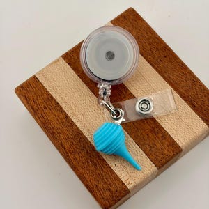 Puede incluir: Un carrete retráctil transparente para tarjetas de identificación con un dije en forma de bulbo azul claro. El carrete tiene una correa de plástico transparente y un clip de metal. El dije es de color turquesa vibrante y tiene una textura acanalada. El carrete está sobre una superficie de madera.